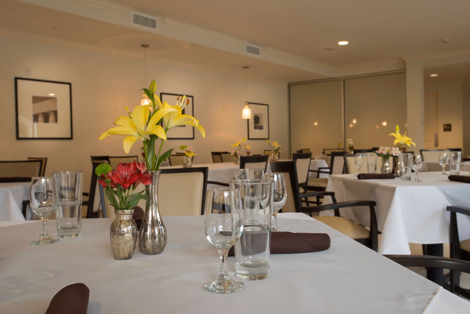 A dining room with tables covered in white tablecloths, each set with glassware, brown napkins, and small vases holding yellow and red flowers. The room has beige walls with framed black and white photographs and pendant lights hanging from the ceiling.
