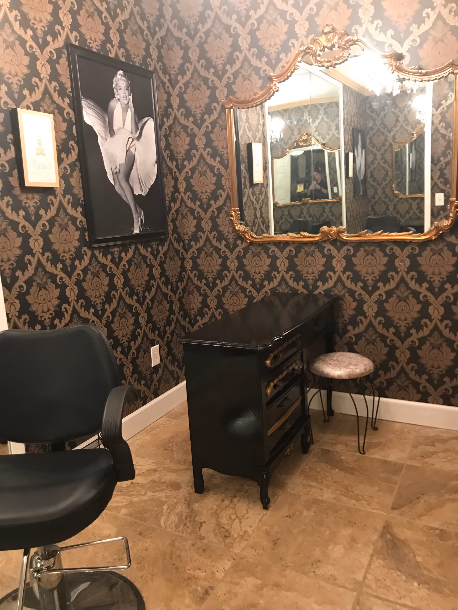Decorative interior room with a styling chair, black vanity, ornate gold-framed mirror, and a Marilyn Monroe portrait on patterned wallpaper.