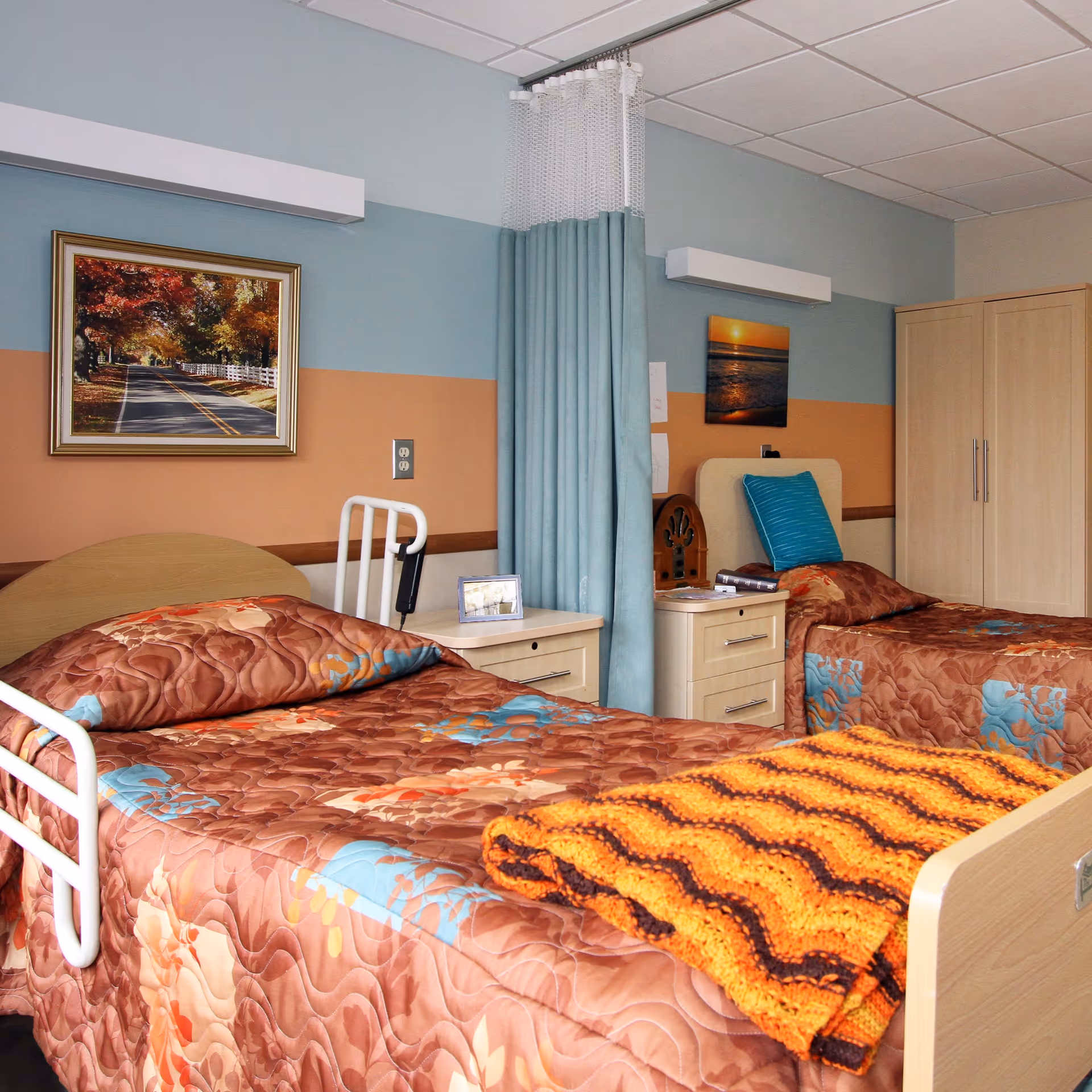 A shared bedroom in a senior living facility with two single beds. Each bed has a brown floral quilt and pillows, with one bed featuring an orange and brown knitted blanket. There is a light blue privacy curtain between the beds. The walls are painted in blue and peach horizontal stripes, decorated with framed pictures. A wooden nightstand with drawers sits between the beds, holding a small framed photo and some books. A wooden wardrobe is visible in the background.