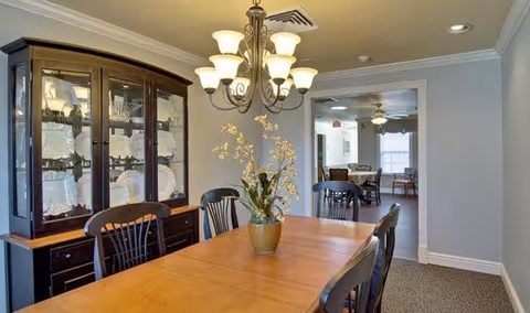 A dining room with a wooden table and six chairs, a chandelier overhead, and a glass-fronted china cabinet filled with dishes. There is a potted plant with flowers on the table. Through an open doorway, another room with additional seating and tables is visible.