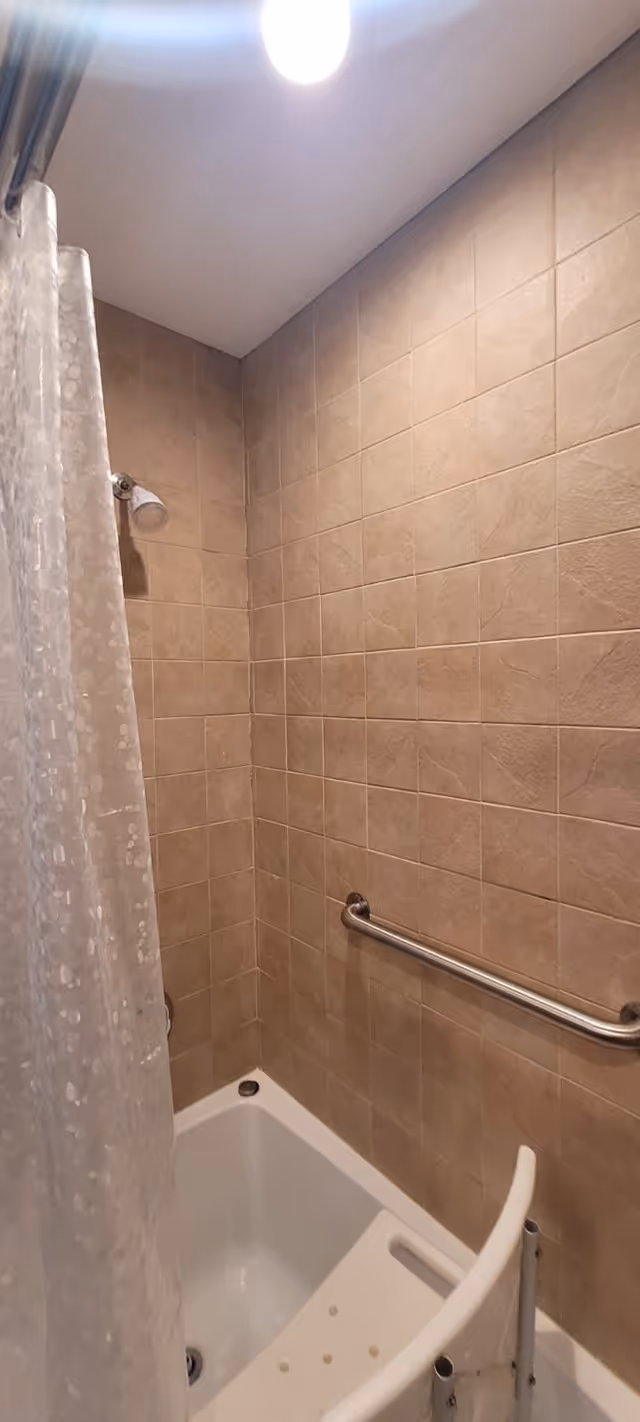 A shower area with beige tiled walls, a white bathtub, a white shower chair, a metal grab bar on the wall, and a translucent shower curtain with a circular pattern. A ceiling light is visible above.