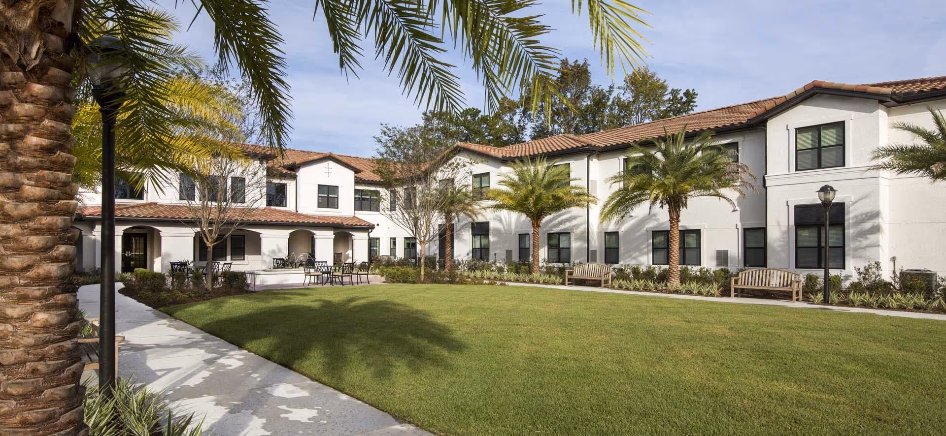 Two-story white building with a central grassy courtyard, palm trees, benches, and outdoor seating.
