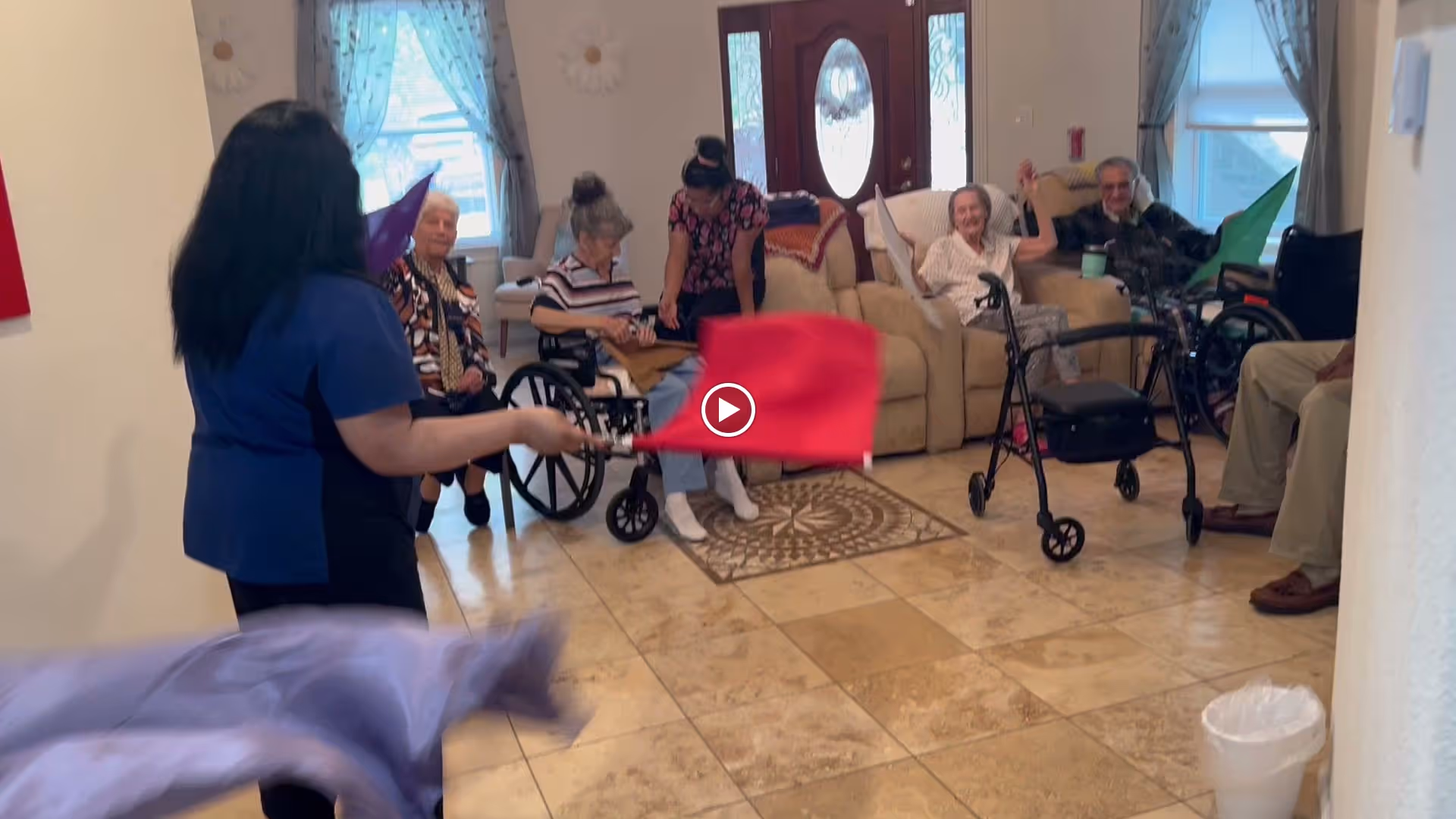 A group of elderly residents and a caregiver in a senior living facility common area. The caregiver is holding colorful flags and engaging with the seated residents, some of whom are in wheelchairs or using walkers. The room has tiled floors, beige walls, and large windows with curtains, creating a bright and welcoming atmosphere.