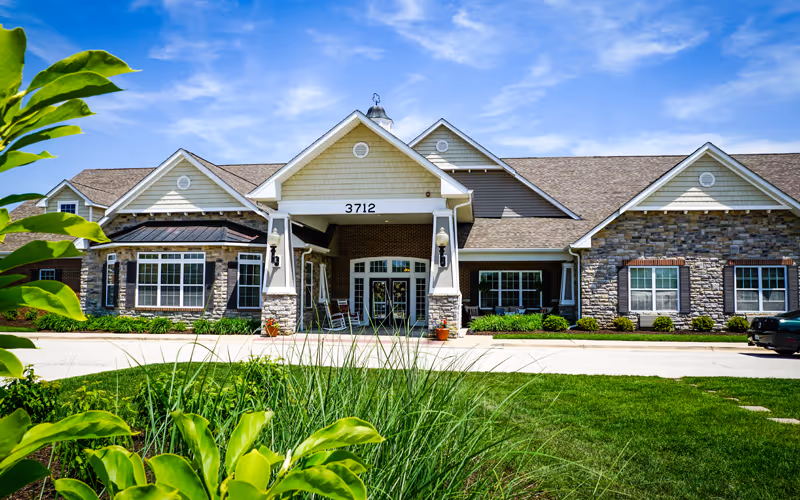 Front exterior view of a single-story senior living facility building with a peaked roof, stone and siding facade, large windows, and a covered entrance with the number 3712 above it. Green shrubs and grass are in the foreground under a bright blue sky.