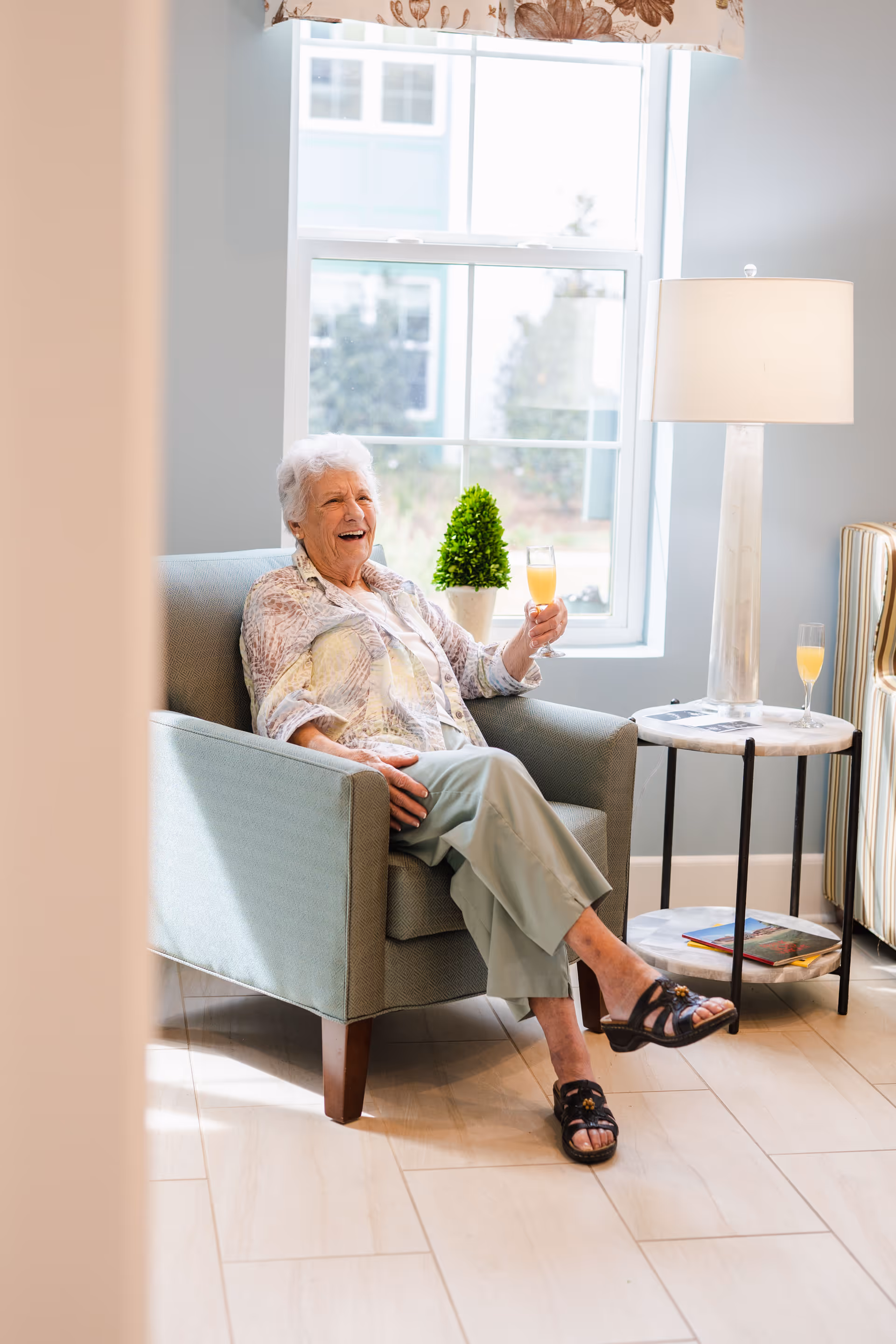 An elderly woman with white hair is sitting comfortably in a light blue armchair near a window, holding a glass of orange juice and smiling. Next to her is a round side table with a lamp, another glass of orange juice, and some magazines. The room has light-colored walls and floor tiles, with natural light coming through the window.