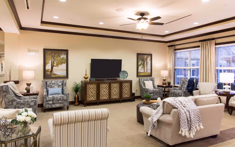 A cozy living room in a senior living facility with multiple upholstered armchairs and sofas arranged around a central coffee table. The room features a flat-screen TV on a decorative wooden cabinet, two framed landscape paintings on the wall, several table lamps, and large windows with curtains letting in natural light.