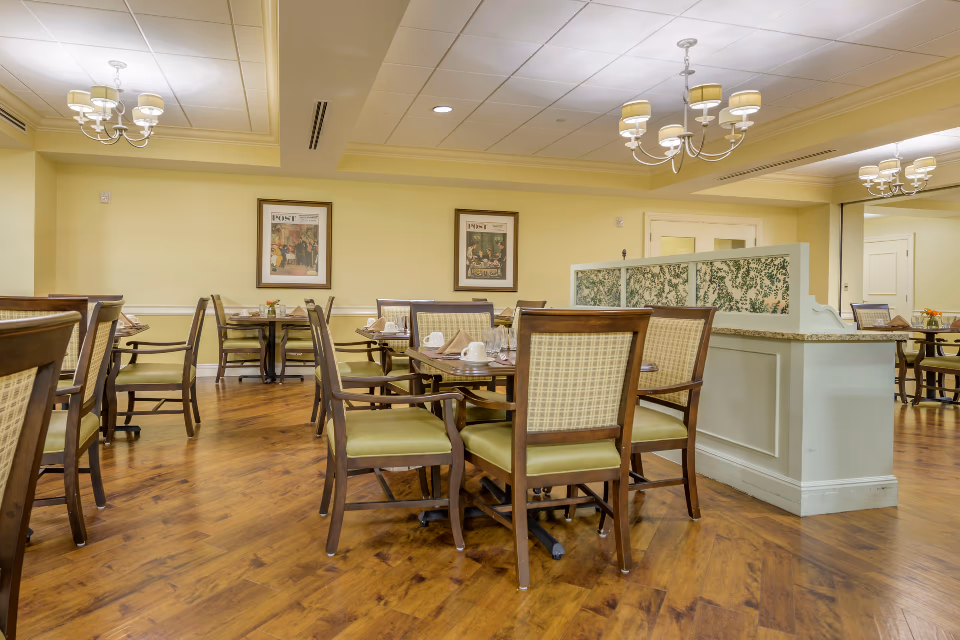 A dining room with wooden floors and several tables and chairs arranged neatly. The chairs have green cushions and patterned backs. The walls are painted light yellow and decorated with framed artwork. There are chandeliers hanging from the ceiling providing warm lighting. A partial divider with a floral design separates part of the room.