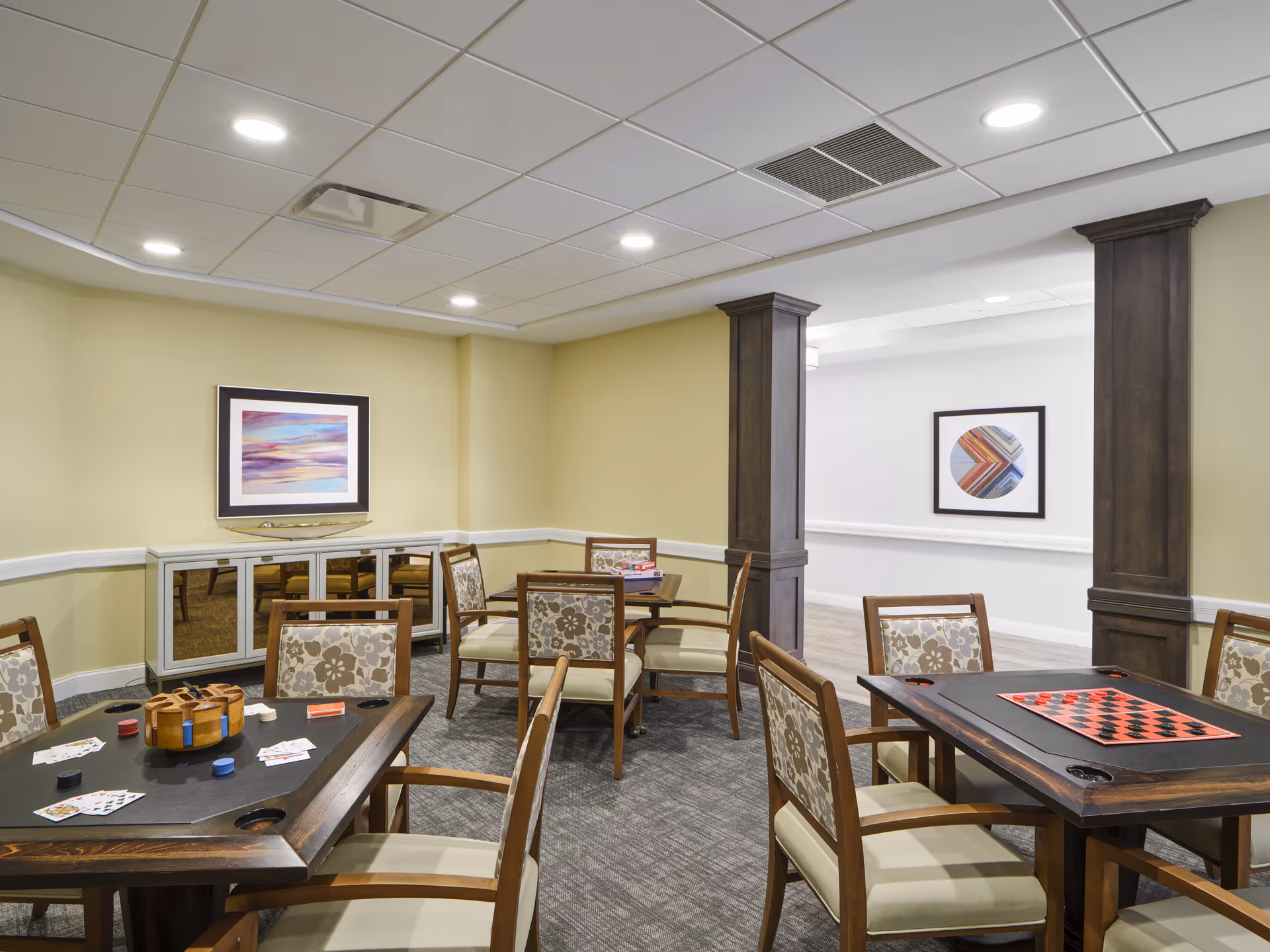 Communal activity room with multiple wooden tables and chairs set up for games like cards and checkers.