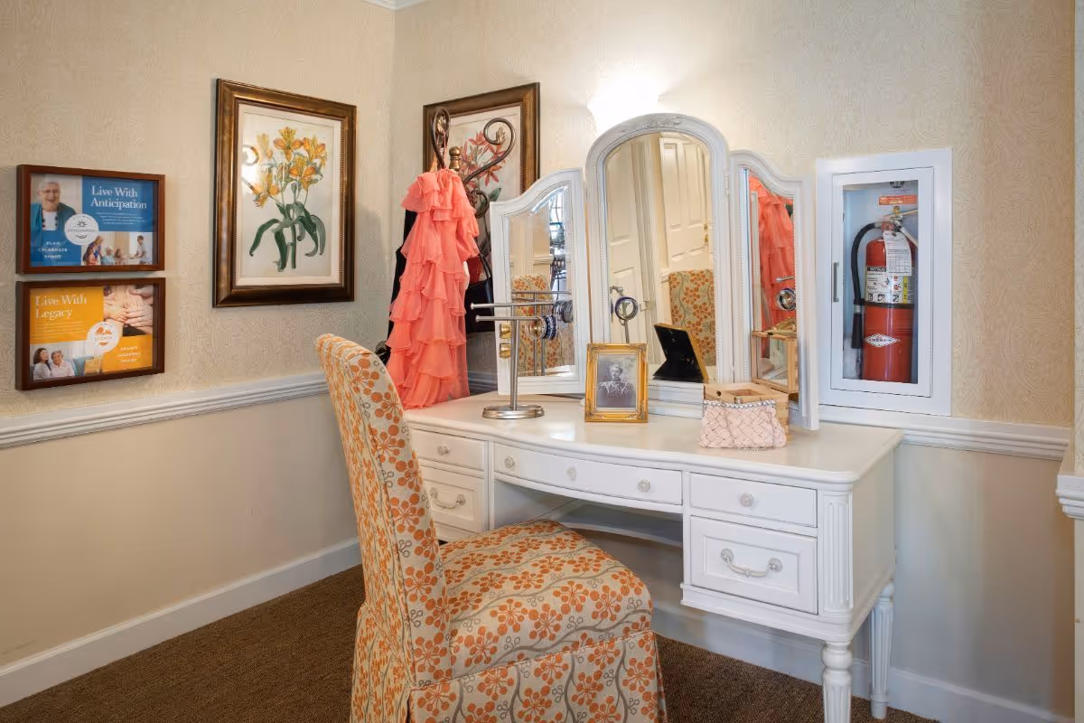 A white dressing vanity with a tri-fold mirror, floral upholstered chair, and decorative items in a softly lit interior corner.