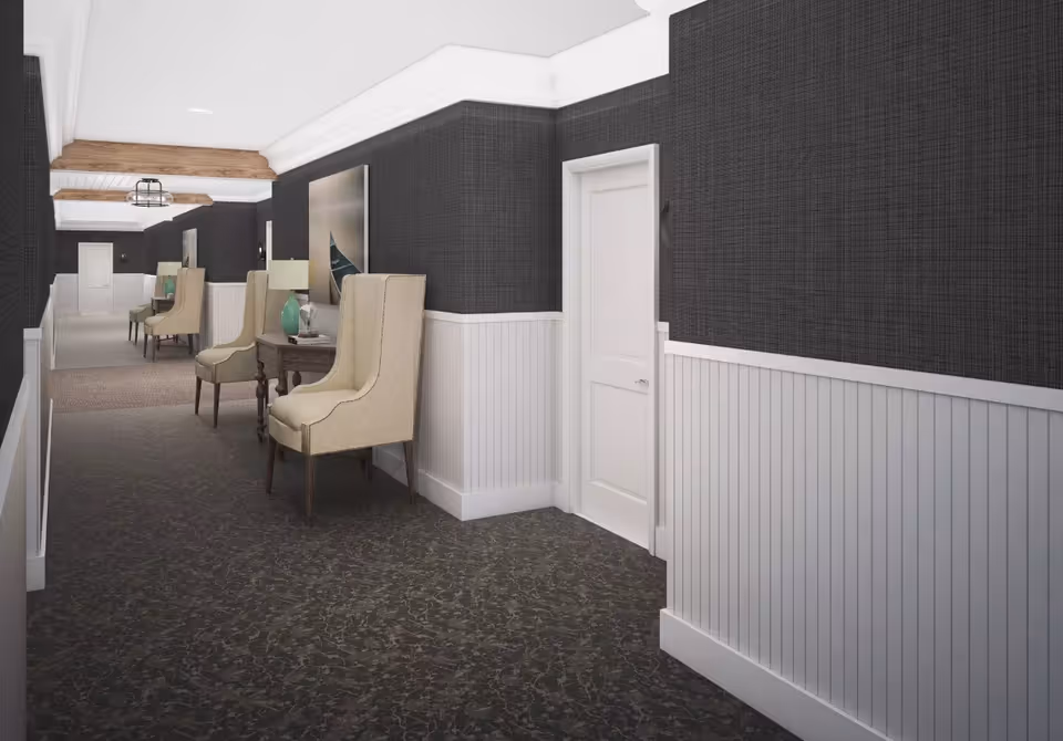 A long hallway with dark textured wallpaper on the upper half of the walls and white wainscoting on the lower half. The carpet is dark with a subtle pattern. Along the hallway, there are beige upholstered chairs paired with small wooden tables holding lamps and decorative items. The ceiling has white crown molding and exposed wooden beams, with modern light fixtures.