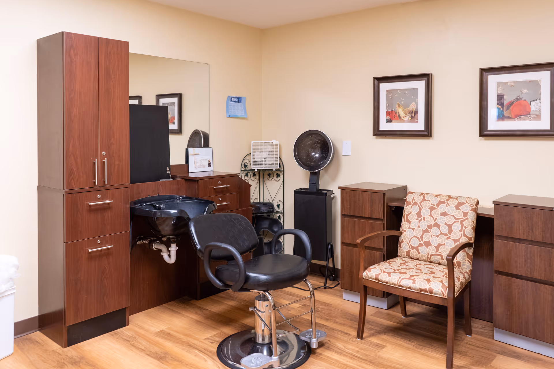 Interior of a salon area in a senior living facility with a black salon chair in front of a hair washing sink, wooden cabinets, a hair dryer, a patterned armchair, and framed artwork on the beige walls.