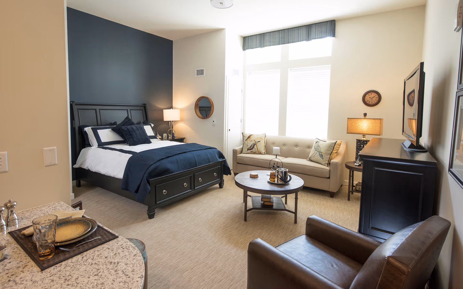 Well-lit studio room with a bed against a dark accent wall, a sofa and coffee table by large windows, a TV on a dresser, and a small kitchenette countertop in the foreground.