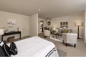 A spacious and well-lit bedroom with a large bed in the foreground, featuring black and white bedding. The room includes a sitting area with two armchairs, a coffee table, and a floor lamp. A console table with decorative items and artwork is positioned against the wall near the door. The overall decor is modern and neutral-toned.