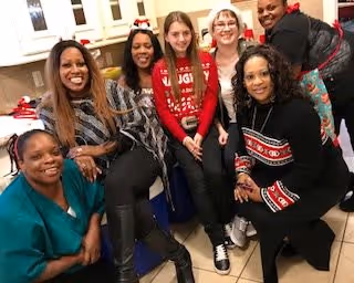 A group of six women smiling and posing together in a kitchen area. The background shows kitchen cabinets, a countertop, and a sink. The women are casually dressed and appear to be enjoying each other's company.