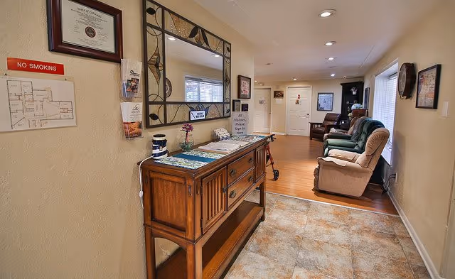 Hallway entry of an assisted living facility with a wooden console table and mirror, tile floor leading to a seating area with recliners.