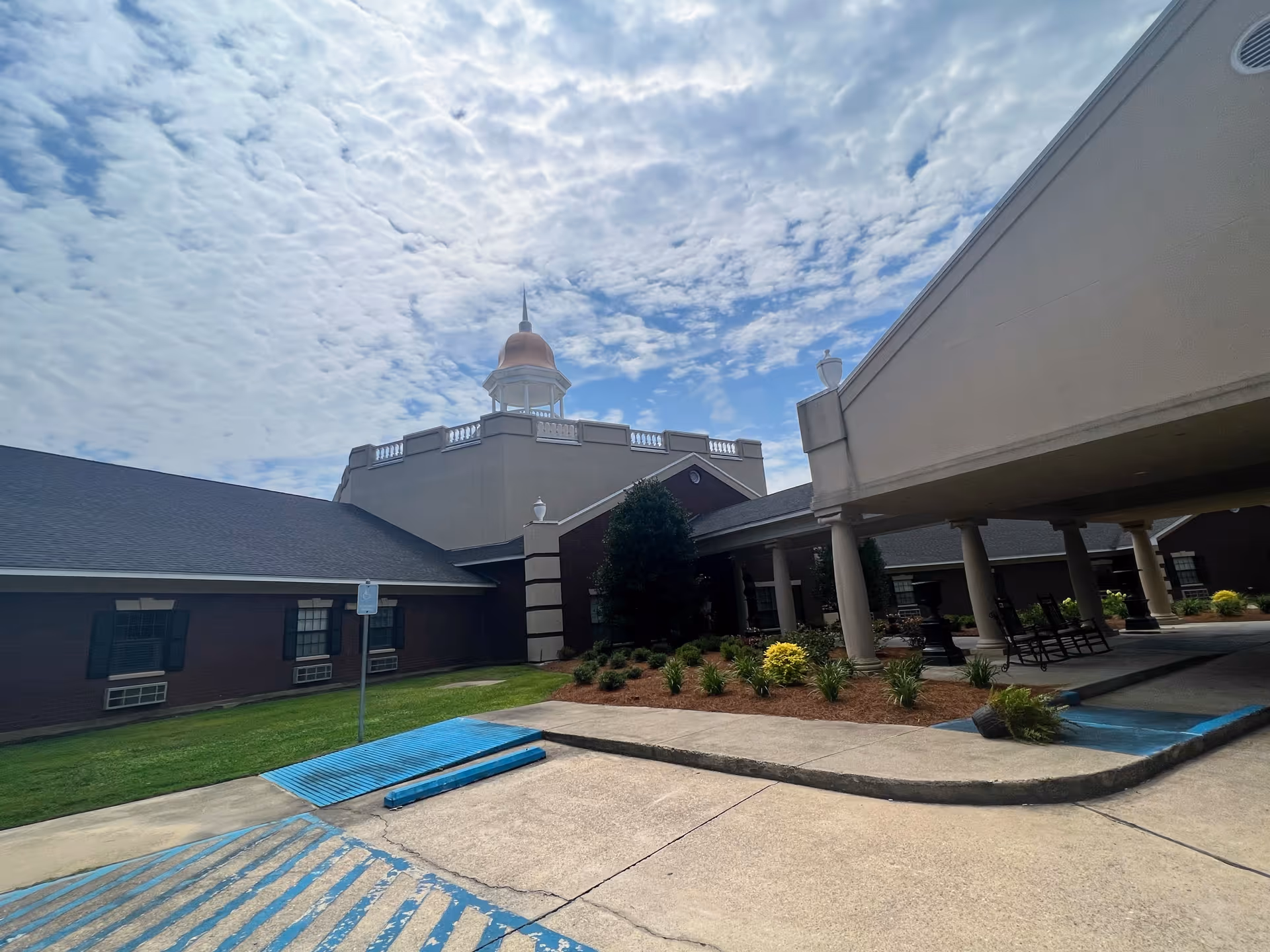 Exterior view of LeGrand Health Care & Rehabilitation Center showing a covered entrance with columns, landscaped garden beds, and a building with a cupola under a partly cloudy sky.