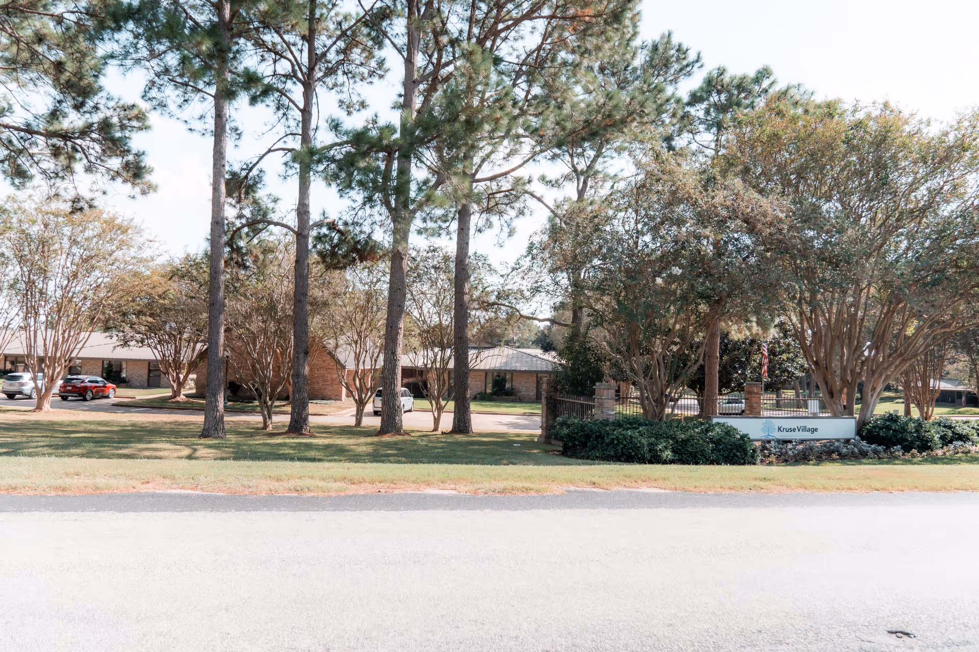 Exterior view of Kruse Village senior living facility showing a single-story brick building partially obscured by tall pine trees and other leafy trees, with a lawn and driveway in front. A sign with the name 'Kruse Village' is visible near the entrance surrounded by shrubs.
