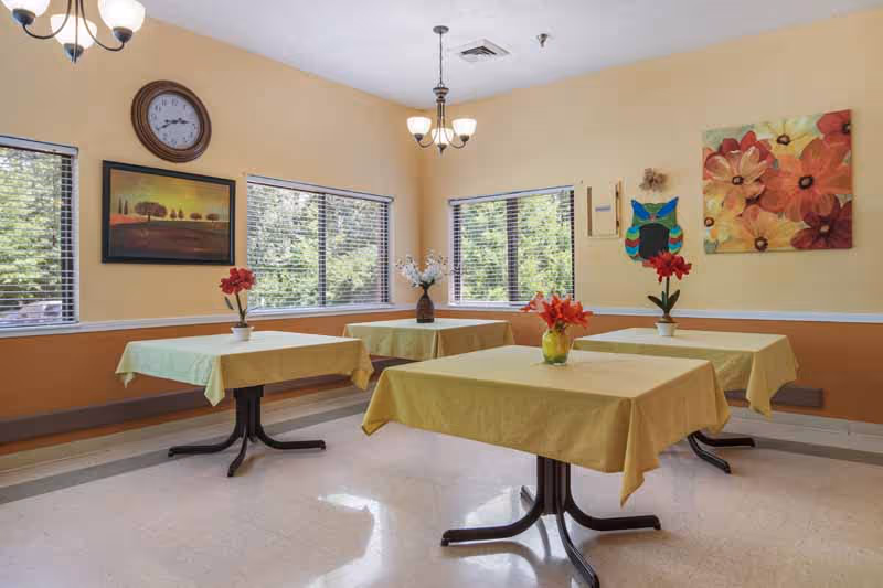 A bright dining room with four square tables covered with yellow tablecloths, each adorned with a vase of colorful flowers. The room has large windows with blinds, light yellow walls, and decorative paintings including a clock and floral artwork. Ceiling lights hang above the tables.