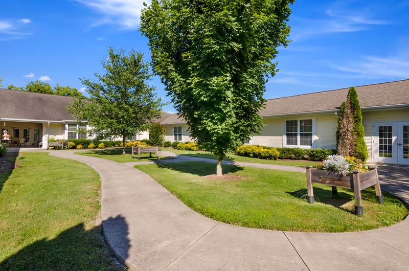 A sunny outdoor courtyard area at American House Bristol with a curved concrete walkway, green grass, trees, shrubs, and a raised wooden planter. The building surrounding the courtyard has beige siding and multiple windows and doors.