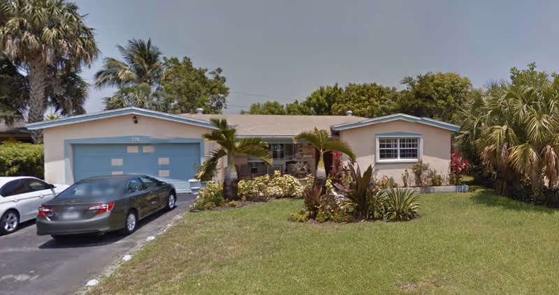 Single-story house with a light-colored exterior and blue trim around the garage door and windows. There are two cars parked in the driveway, a well-maintained lawn, and various plants and palm trees in the front yard under a clear sky.