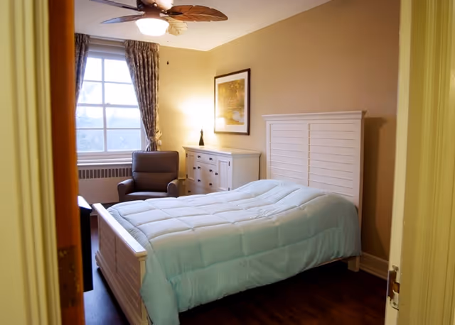 A cozy bedroom with a white wooden bed frame and a light blue comforter. There is a gray armchair near a window with patterned curtains, a white dresser with a lamp and a framed picture above it. The room has beige walls and wooden flooring.
