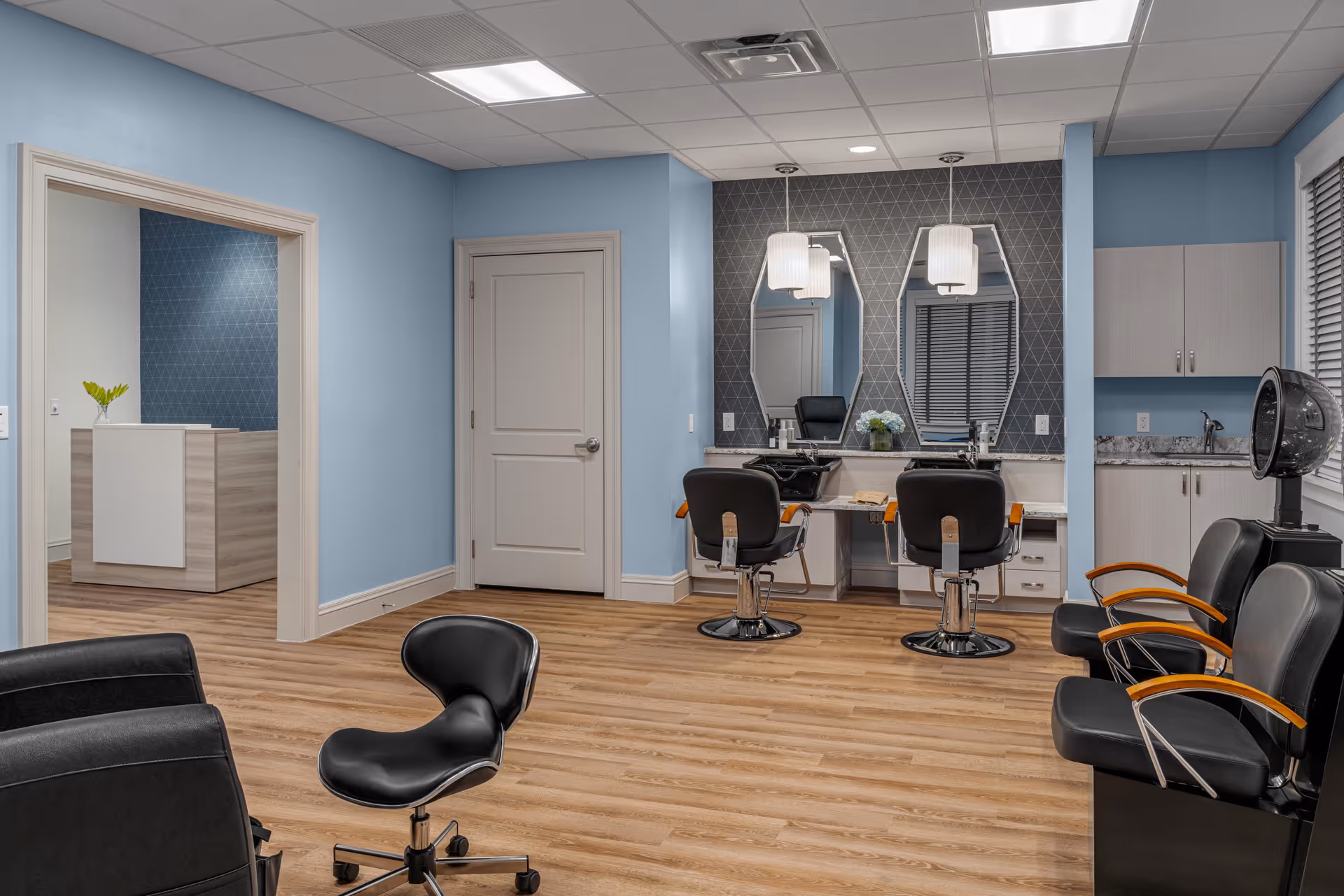 Interior view of a salon area in a senior living facility with two styling chairs in front of mirrors, a hair dryer chair, and additional seating. The walls are painted light blue, and the floor has wood-like finish. There is a reception desk visible through an open doorway.
