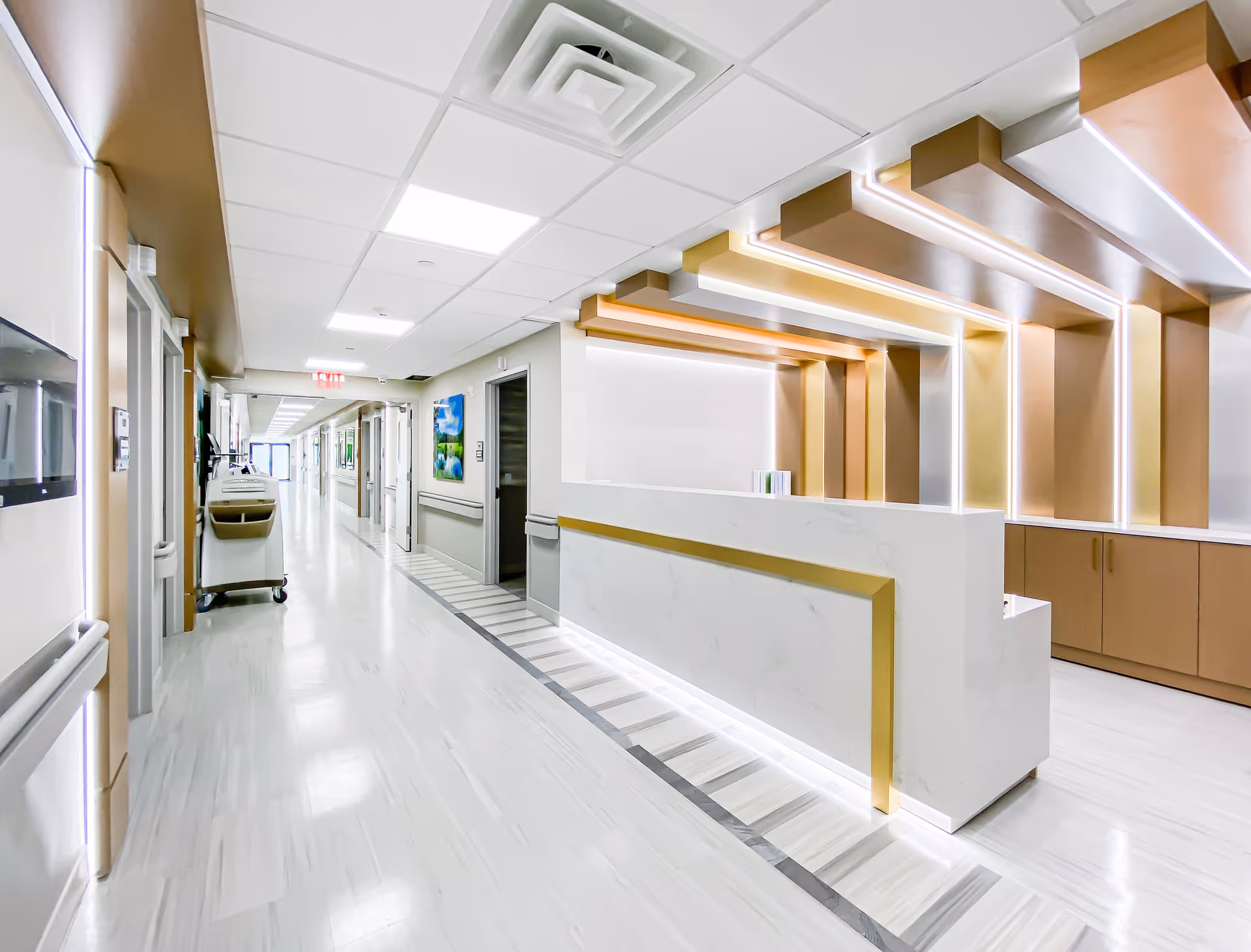 Bright and modern hallway in a senior living facility featuring a sleek white reception desk with gold accents and illuminated ceiling design. The corridor extends into the distance with doors on either side and a cleaning cart visible along the left wall.