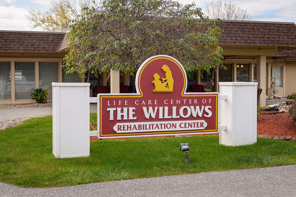 Outdoor view of the entrance sign for Life Care Center of The Willows Rehabilitation Center, with a building and landscaping in the background.
