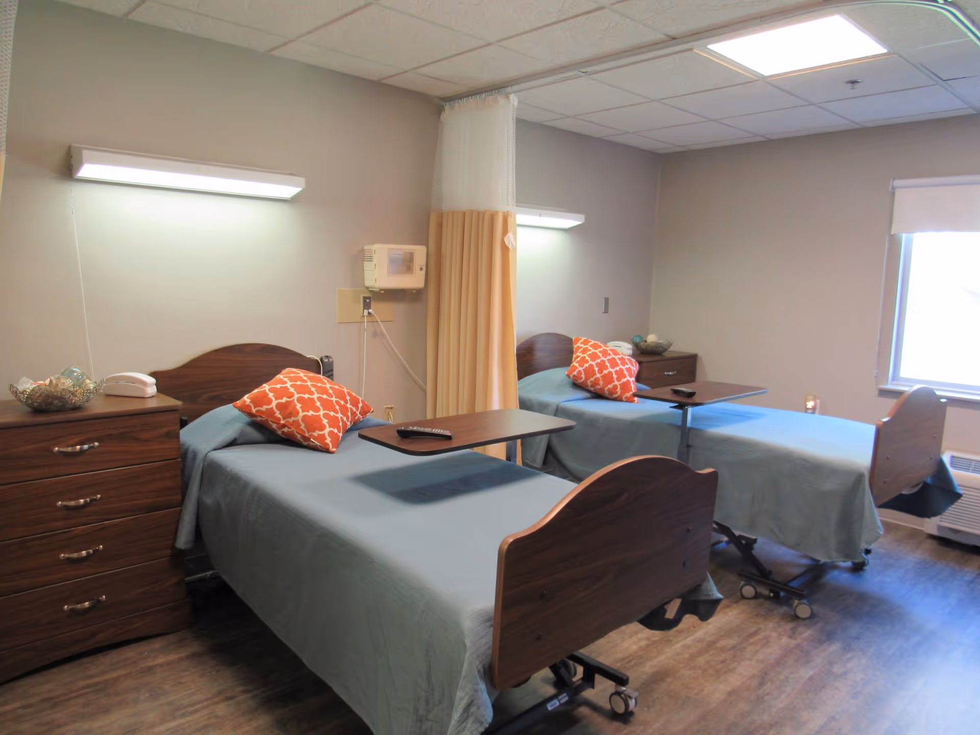 A nursing facility room with two hospital-style beds, each with a blue bedspread and an orange patterned pillow. Each bed has an overbed table with a remote control on it. There is a wooden nightstand with drawers between the beds and a window with a white blind on the right side. The room has wood-look flooring and neutral-colored walls with overhead fluorescent lighting.