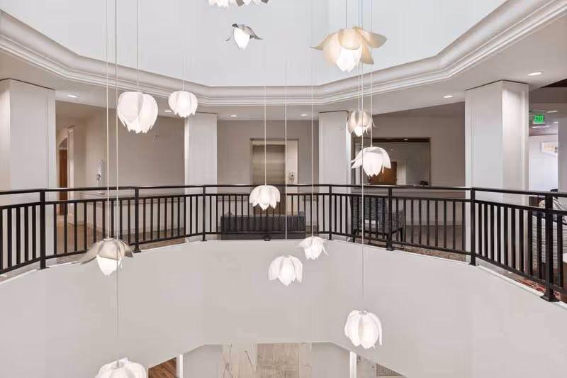 Interior view of a senior living facility featuring a circular balcony with black railings overlooking a lower floor. Several white flower-shaped pendant lights hang from the ceiling at varying heights. The walls are painted white, and there is an elevator and seating area visible in the background.