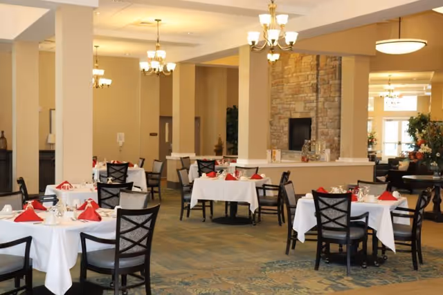 Dining room with multiple tables set with white tablecloths and red napkins, chandeliers, and a stone fireplace.