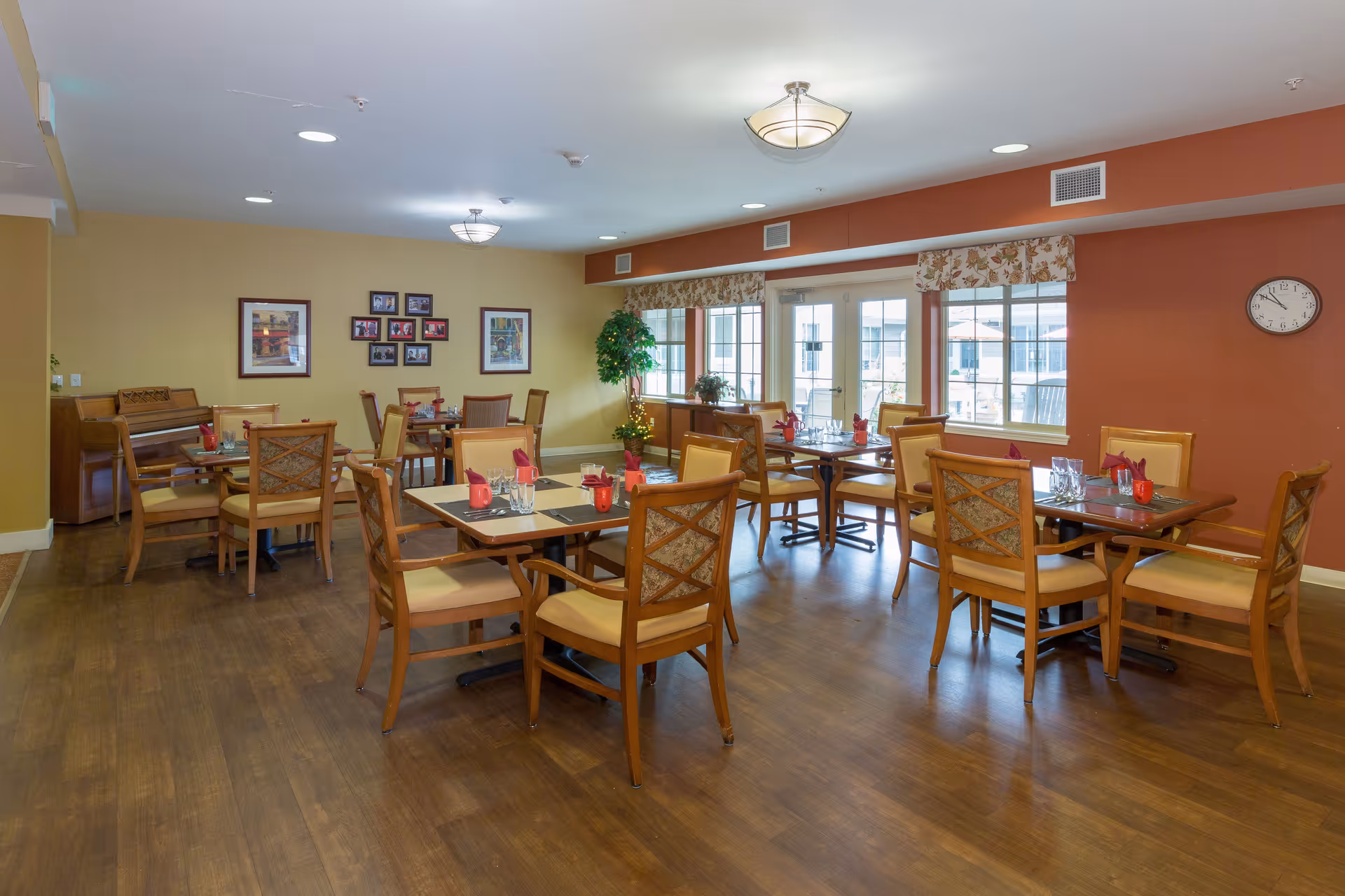A dining room with several wooden tables and chairs arranged neatly. Each table is set with placemats, glasses, and red napkins. The room has wooden flooring, large windows with floral valances, framed pictures on the walls, a piano in the corner, and ceiling lights providing illumination.