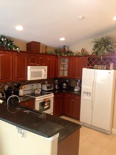 A kitchen with dark wood cabinets, a white microwave above a white stove, a white refrigerator with water and ice dispenser, and a black countertop with a sink. There are various kitchen appliances on the counter and decorative greenery on top of the cabinets.