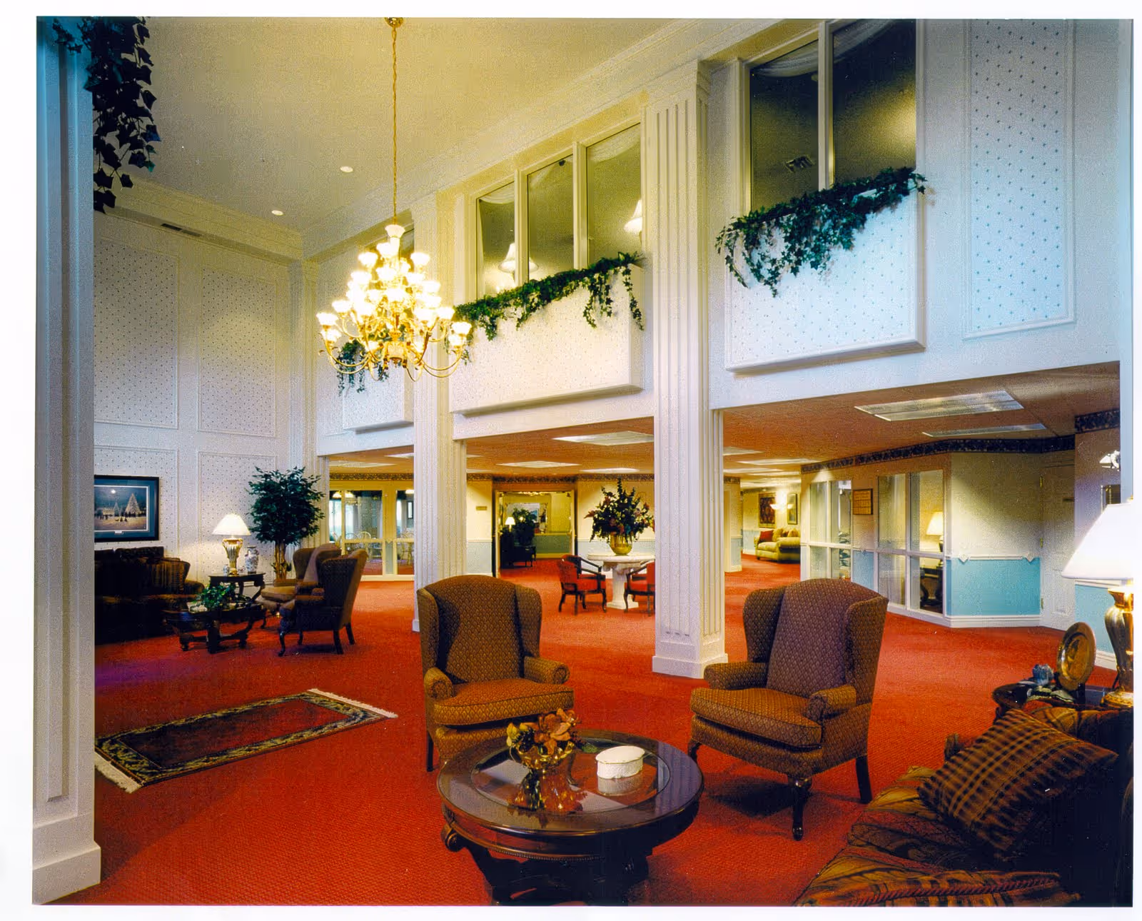 A spacious and well-lit senior living facility common area with red carpet flooring, multiple seating arrangements including armchairs and sofas, a round glass coffee table with decorative items, a large chandelier hanging from the ceiling, and greenery decorating the upper wall ledges.
