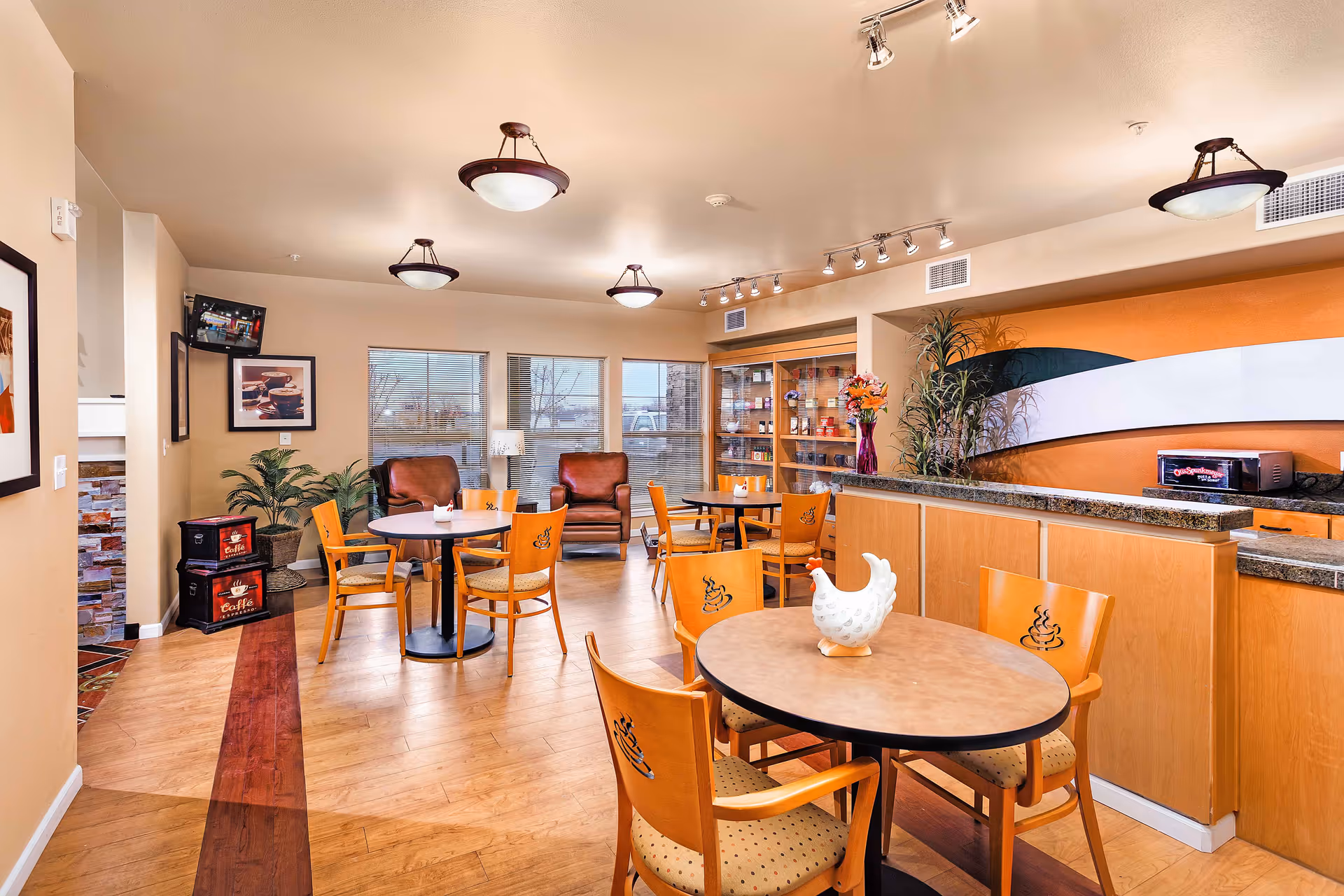 A bright and cozy dining area in a senior living facility with round tables and wooden chairs featuring a coffee cup design on the backrests. The room has large windows letting in natural light, two comfortable armchairs near the windows, a countertop with a microwave, and decorative plants and artwork on the walls.