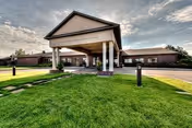 Front entrance of a single-story senior living facility with a covered porte-cochere, columns, and a well-manicured lawn under a cloudy sky.