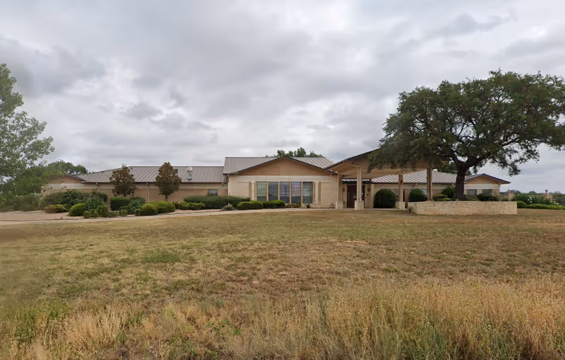Exterior view of a single-story assisted living facility building with beige brick walls and a metal roof. The building is surrounded by trimmed bushes and trees, with a large grassy area in the foreground under a cloudy sky.