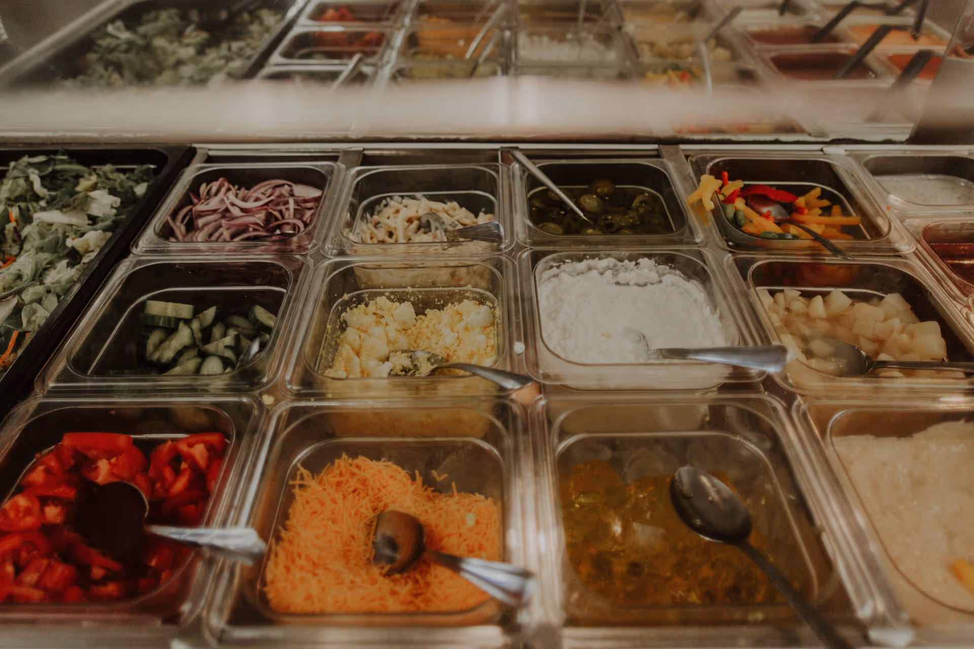 Buffet-style salad bar with multiple clear trays holding assorted salad toppings and condiments.