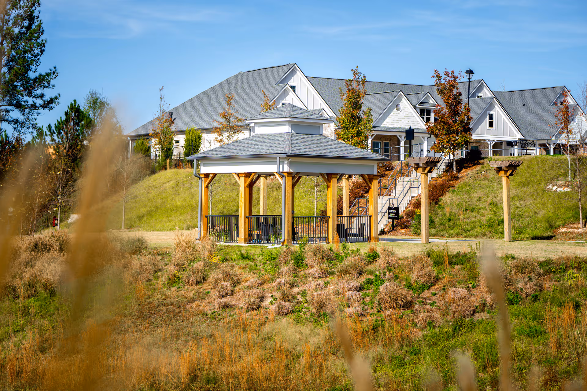 Outdoor view of The Mansions at Gwinnett Park Senior Independent Living featuring a wooden gazebo with seating, surrounded by grass and plants, with a large building and stairs in the background under a clear blue sky.
