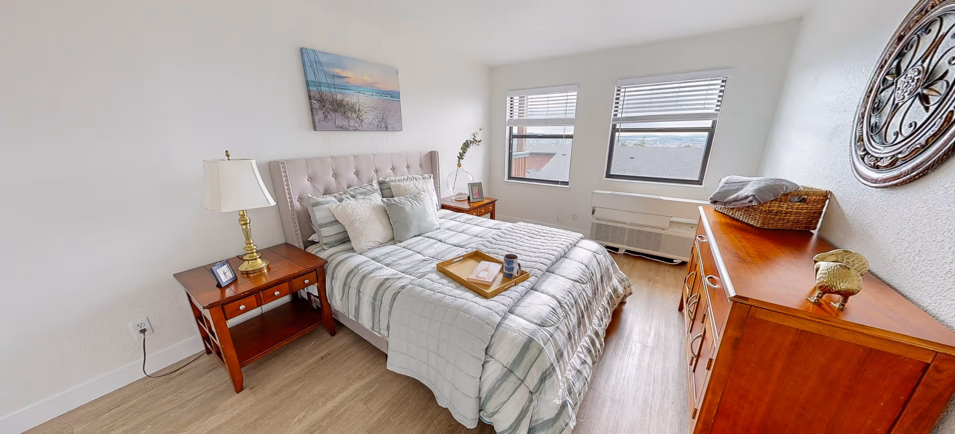 A bright and cozy bedroom with a neatly made bed featuring a tufted headboard and multiple pillows. There is a wooden nightstand with a lamp and a small framed photo on the left side of the bed. On the right side, there is another wooden nightstand with a glass vase holding a branch. Two windows with blinds allow natural light to fill the room. A wooden dresser with decorative items and a basket is positioned against the right wall, which also has a round metal wall decoration. The floor is light wood, and a tray with a book and a mug is placed on the bed.