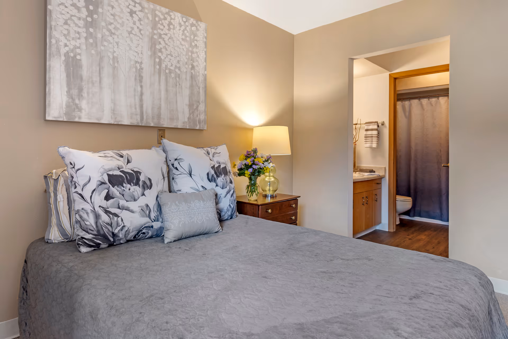 A neatly made bedroom with a gray bedspread, floral pillows, a bedside table with lamp and flowers, and an open doorway leading to a bathroom.
