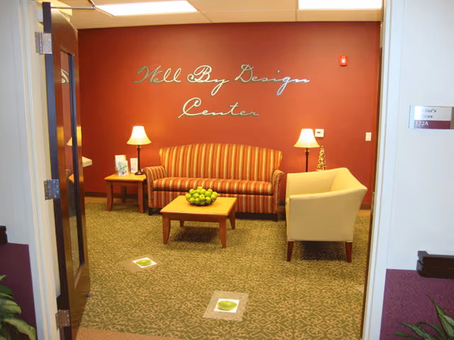 A cozy sitting area with a striped sofa, a beige armchair, two side tables with lamps, and a coffee table with a bowl of green apples. The wall behind the sofa is painted red and has the words 'Well By Design Center' written on it in cursive. The floor is carpeted with a patterned design.