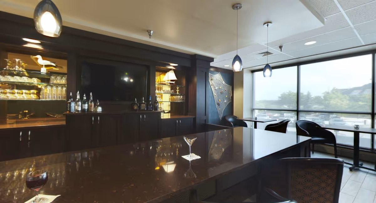 Interior view of a lounge area with a dark wood bar counter, glass shelves with glassware and bottles, a flat screen TV mounted on the wall, and several tables and chairs near large windows letting in natural light.