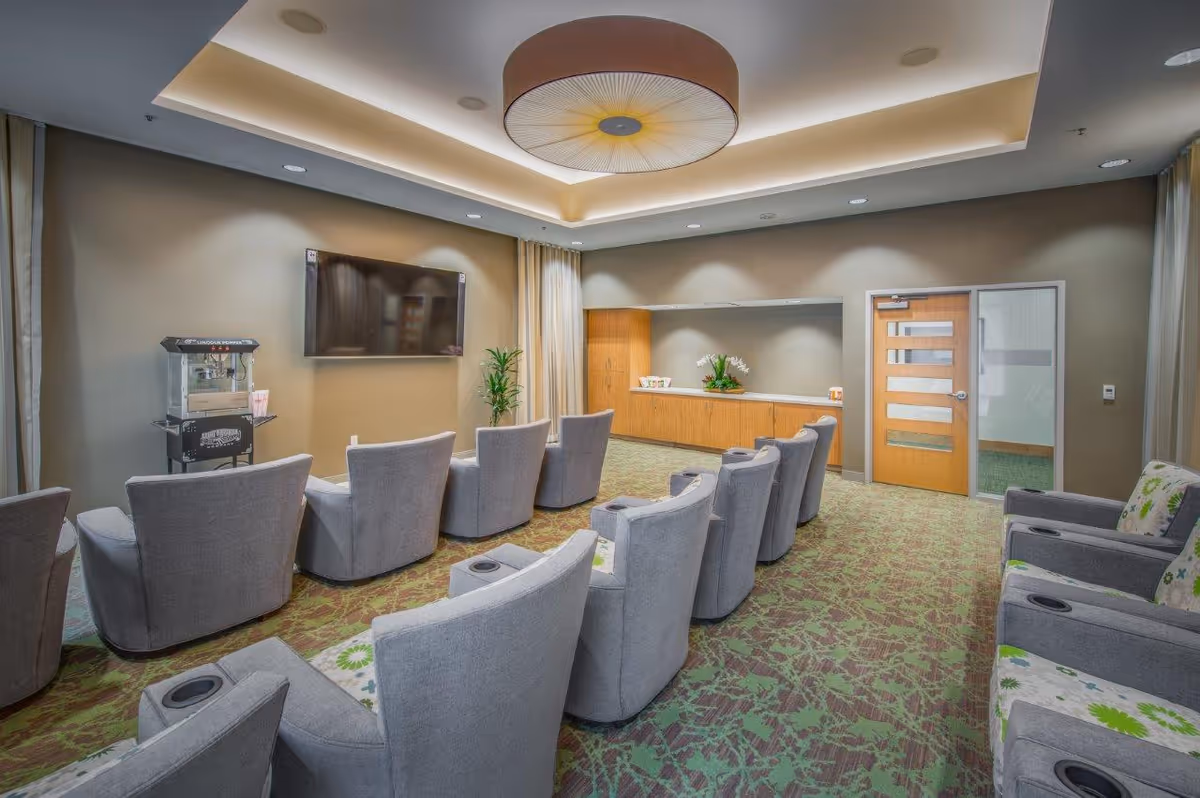 A modern lounge area with two rows of gray upholstered chairs facing a wall-mounted flat screen TV. The room has a green patterned carpet, a popcorn machine on a stand, a wooden cabinet with decorative plants, and a wooden door with glass panels. The ceiling features recessed lighting and a large circular light fixture.