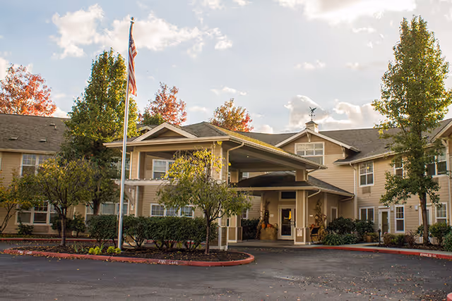 Front entrance of a senior living building with a covered drive-up canopy, landscaping, and an American flag.