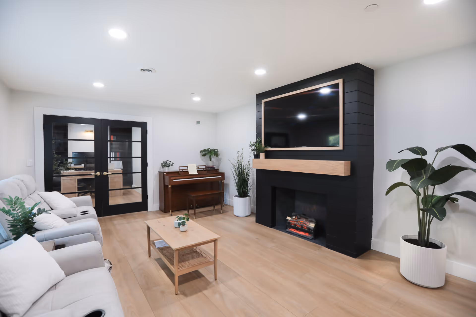 A modern living room with light wood flooring, a black fireplace with a mounted flat-screen TV above it, a wooden coffee table with small plants, a piano against the back wall, and two large potted plants. There are two light-colored recliner sofas on the left side and black double doors with glass panels in the background.