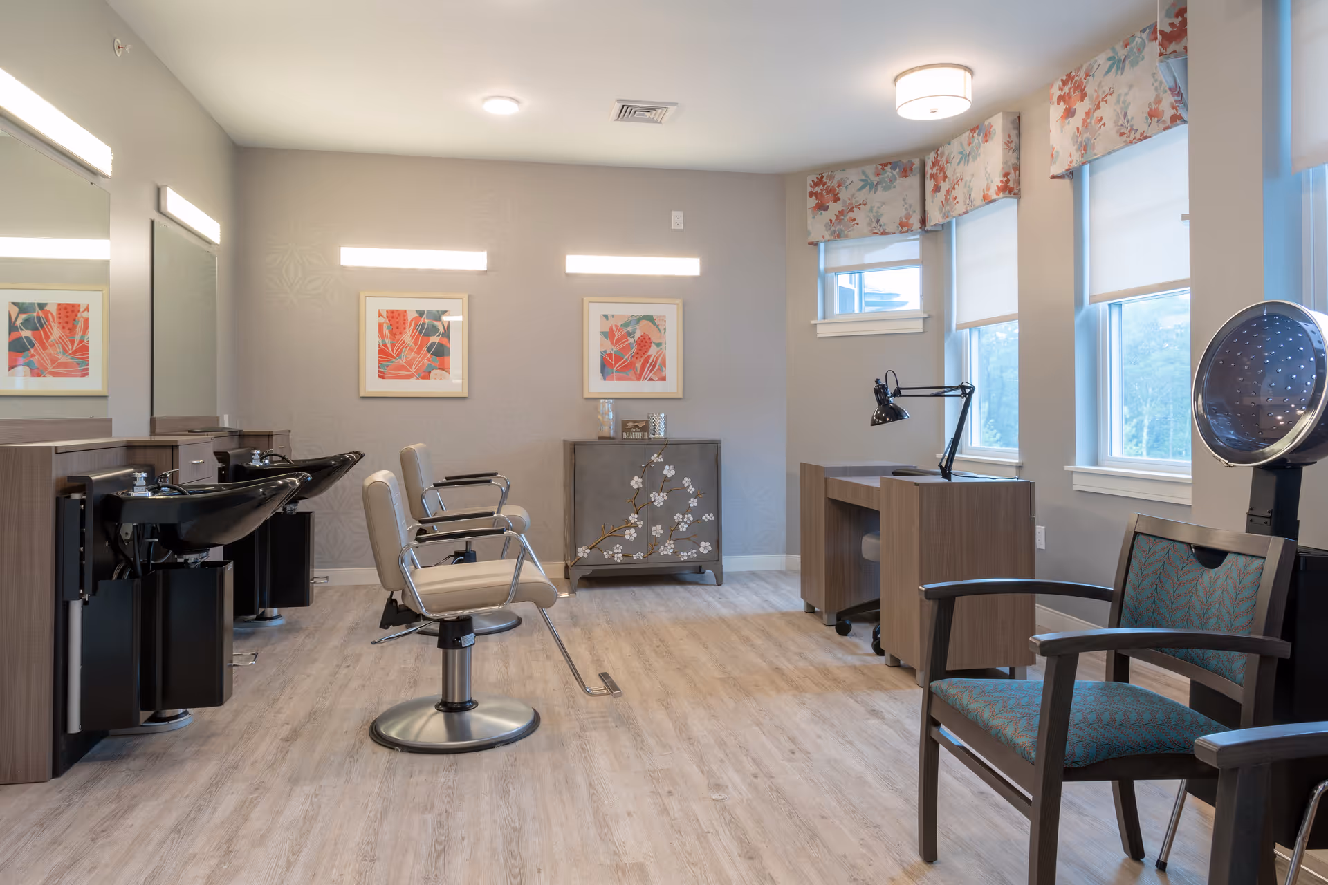 A bright and clean hair salon area with two styling chairs in front of black wash basins on the left, a wooden desk with a black adjustable lamp near windows with floral valances on the right, and a hair dryer chair in the foreground. The walls are light gray with two colorful framed artworks and a decorative cabinet with a floral design.