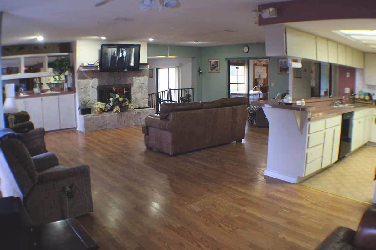 Open communal living area with sofas and armchairs facing a stone fireplace with a mounted TV and an adjacent kitchen with a breakfast bar.