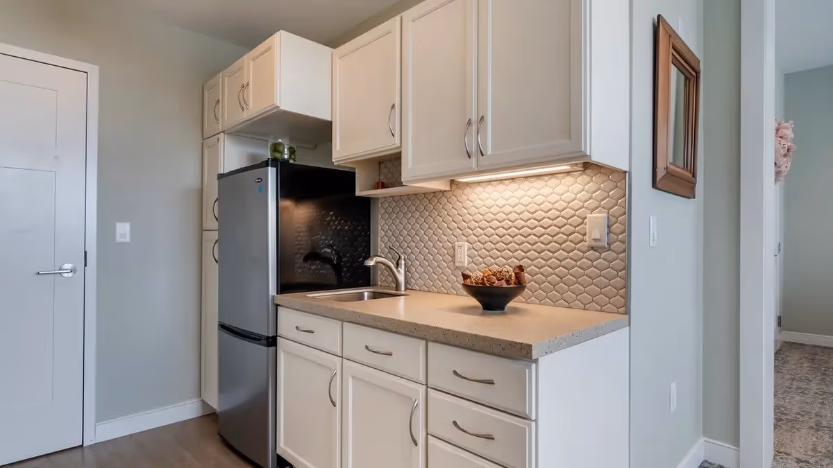 A modern kitchen area with white cabinets, a stainless steel refrigerator, a sink, and a beige countertop. The backsplash features a textured tile design, and there is a bowl with decorative items on the counter. The kitchen is well-lit with under-cabinet lighting.