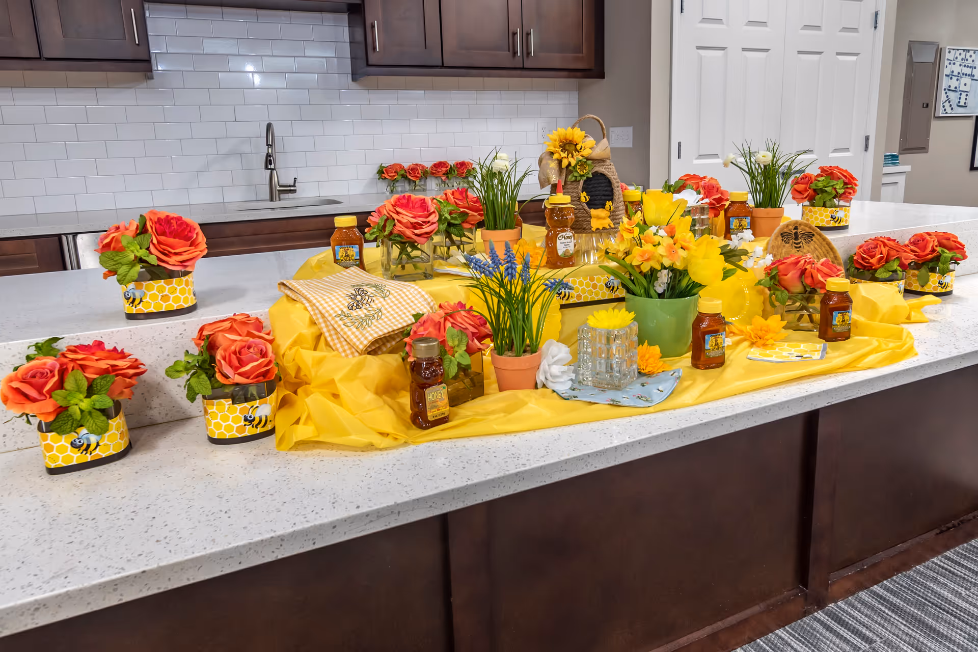 A kitchen counter decorated with a yellow cloth, various potted flowers including red roses and yellow daffodils, small jars of honey, and bee-themed decorations. The background shows dark wood cabinets, a white subway tile backsplash, and a stainless steel sink with a faucet.